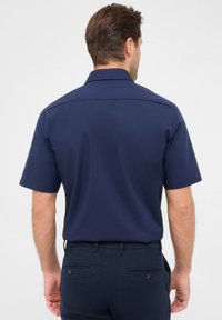 Marineblaues Kurzarmhemd mit geradem Schnitt und Rückeinsatz, aus einem glatten Stoff gefertigt. Getragen mit dunklen Hosen, die über Gesäßtaschen verfügen.