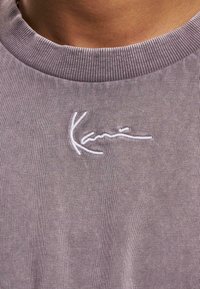 Pilkas sportinis megztinis, turintis nėriniuotą apykaklę ir balta siuvinėta logo. Medžiaga minkšta ir tekstūringa, su prigludusia forma.