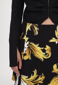 Femme portant un haut noir à manches longues et une jupe noire avec de grands motifs floraux jaunes et blancs, laissant apparaître une partie de son ventre.