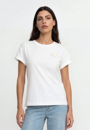 Kobieta o długich ciemnych włosach, ubrana w białą koszulkę z podwiniętymi rękawami z wzorem stokrotki oraz jasnoniebieskie jeansy, stojąca na białym tle.