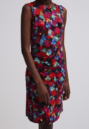 Femme portant une robe sans manches, longueur genou, à motif géométrique carré audacieux rouge, bleu et multicolore, posant devant un fond uni.