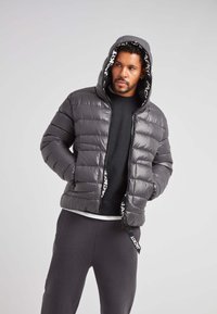 Chaqueta de plumas gris con capucha, con un acabado brillante y costuras horizontales. Acentuada con una cinta de logo negra en el interior de la capucha.