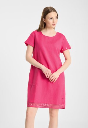 Vrouw draagt een roze jurk met korte mouwen en knielengte met een kanten rand aan de zoom, staat met gevouwen handen en kijkt opzij.