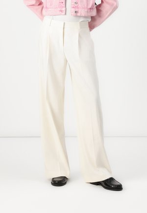 Personne portant un pantalon blanc à jambes larges, des mocassins noirs et une veste courte rose, les mains derrière le dos.