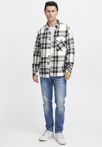 Una camicia flanella a quadri in bianco e nero con taschino sul petto, abbinata a jeans blu sbiaditi e scarpe da ginnastica bianche, in piedi contro uno sfondo semplice.