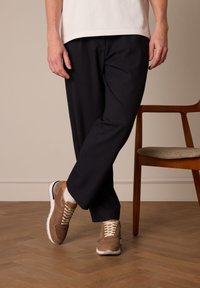 Bruine suède sneakers met crèmekleurige veters, een witte zool en tan accenten. Draag ze met zwarte op maat gemaakte broeken, staand op een houten vloer nabij een stoel.