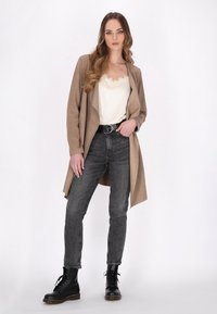 Lang beige blazer over en hvid top med blondekanter, parret med højtaljede sorte jeans og sorte snørebåndsstøvler, sat mod en lys baggrund.