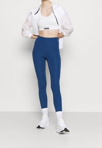 Modré legíny s vysokým pasem a hladkým povrchem, kombinované s bílým zkráceným sportovním topem a lehkou průhlednou bundou.