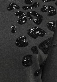 Mørk grå stoff med svarte paljetter i leopardmønster; paljettene skaper en teksturert, glitrende kontrast mot det myke bakgrunnsmaterialet.