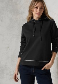 Zwarte hoodie van zachte stof, met een trekkoord, witte bies langs de zoom en mouwen, en een relaxte pasvorm. Draag met donkere jeans.
