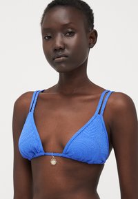 Plavi bikini top u obliku trokuta s teksturiranim materijalom i složenim uzorcima, tankim trakama i zlatnim detaljem u obliku sunca u središtu.