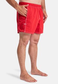 Red swim shorts with elastic waistband, side pockets, and white stitching details; lightweight, quick-drying material.