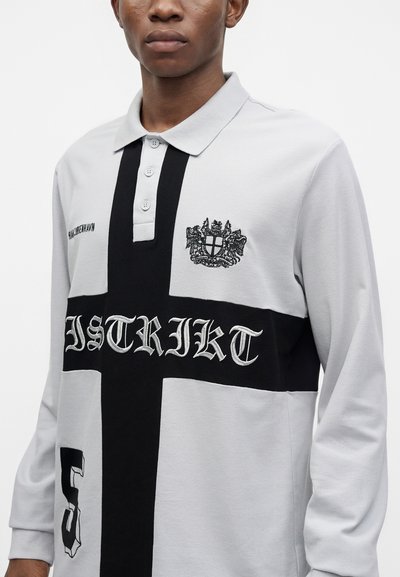 Homme portant un polo gris clair à manches longues avec un motif de croix noire, un blason brodé, le texte « DISTRIKT » et le numéro 5 à l'avant.