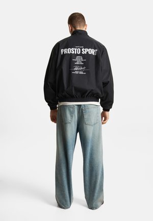 Mężczyzna ubrany w czarną kurtkę Prosto Sport z białym napisem na plecach oraz jasne, szerokie jeansy, stoi tyłem do białego tła.