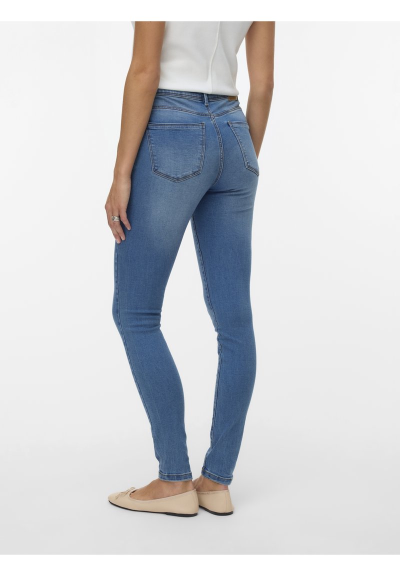 Femme portant un jean skinny bleu clair et des ballerines beiges, debout de dos devant un fond blanc uni.