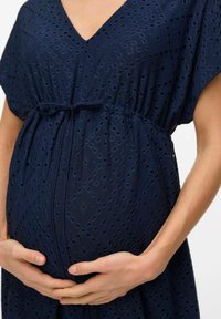Abito premaman blu navy realizzato in tessuto testurizzato con motivi ad occhiello, scollatura a V, e una vita con cordoncino che si adatta su una pancia arrotondata.