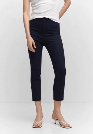 Kobieta w dopasowanych granatowych spodniach 7/8, białym bezrękawnym topie i beżowych sandałach na niskim obcasie stoi na tle jasnego, jednolitego tła.