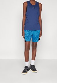 Granatowy bezrękawnik sportowy z małym logo, w połączeniu z turkusowymi szortami oraz czarnymi sneakersami z żółtymi akcentami i białymi skarpetkami.