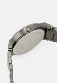 Reloj de acero inoxidable plateado con caja redonda, correa texturizada y una corona lateral con el nombre de la marca. Marcado en la parte posterior como resistente al agua.