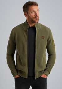 Olivgrüner Zip-Up-Pullover aus strukturiertem Material, hoher Kragen, Seitentaschen und einem Badge-Detail am linken Ärmel. Figurbetonter Schnitt.