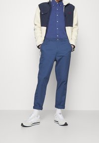 Man med blå knapp-up skjorta, marinblå och krämfärgad fleecejacka, blå byxor med uppvikt nederkant, vita strumpor och vita sneakers, stående med händerna i fickorna.