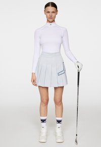 Biała bluzka z długim rękawem, jasnoniebieska spódnica plisowana, białe buty sportowe i skarpetki w wzory z logo. Kij golfowy trzymany w prawej ręce.