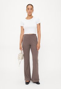 Witte geribbelde crop top met gegolfde randen, gecombineerd met bruine geribbelde flared pants. Houdt een kleine crèmekleurige handtas vast en draagt zwarte schoenen.