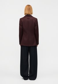 Μακρυνό σκούρο μπουφάν με μία πίσω σκίσιμο και μανσέτες με κουμπιά, σε συνδυασμό με μαύρο παντελόνι με φαρδιά πόδια και ριγέ μοτίβο.