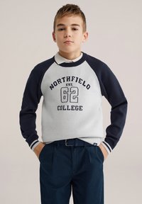 Sweat-shirt gris avec manches raglan bleu foncé, présentant l'inscription "NORTHFIELD EST. 62 COLLEGE" sur le devant. Porté avec un pantalon bleu marine.