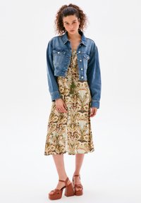 Denimjacke mit Fronttaschen über einem ärmellosen, gemusterten Kleid mit grünen und braunen Blattmotiven; das Model trägt braune Plattformabsätze.