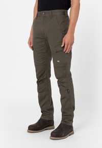Olive green cargo pants with multiple pockets, reinforced knees, and a fitted design, made of durable fabric. Worn with brown work boots.