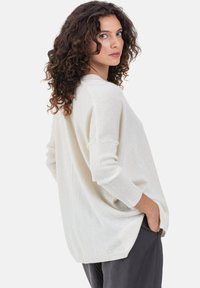 Pull en maille crème avec une coupe décontractée, des poignets côtelés et des fentes latérales. Porté avec un pantalon foncé, il présente une texture douce et un design décontracté.