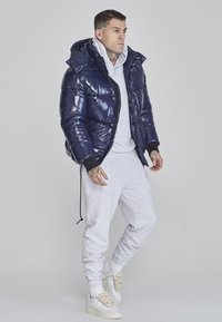 Navy blue puffer jacket with glossy finish, hood, and zip closure. Worn over a gray hoodie and sweatpants. Light-colored sneakers.