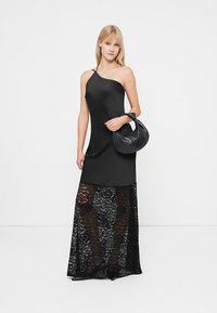 Satin juoda suknelė su viena petnešėle, pereinanti į ilgą nėrinių sijoną. Modelis laiko apvalią juodą rankinę. Lygi tekstūra ir elegantiškas dizainas.