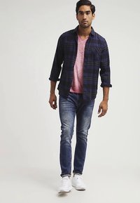 Pehmeä flanellipaita tummansinisessä ruutukuosissa vaaleanpunaisen v-kaula-aukkoinen t-paidan päällä, yhdistetty kapealahkeisiin kuluneen sinisiin farkkuihin ja valkoisiin lenkkareihin.