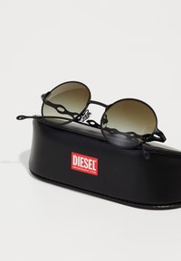 Ochelari de soare rotunzi negri cu lentile gradient, având un design de ramă unic și un toc din piele neagră cu un logo roșu "DIESEL".