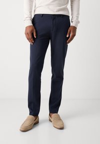 Marineblauwe op maat gemaakte broek met een gladde textuur, slim fit ontwerp en een voornaad. Gecombineerd met lichtgekleurde loafers voor contrast.