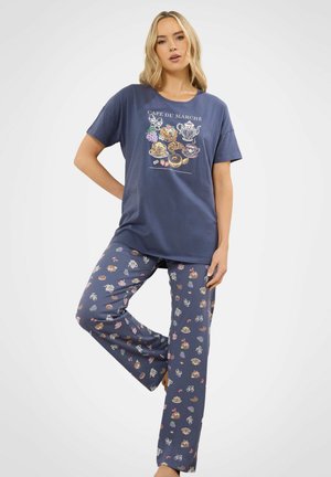 Donna che indossa un completo pigiama blu navy con illustrazioni a tema colazione e la scritta "Café du Marché", in piedi con una gamba piegata.
