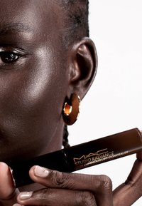 MAC Studio Radiance 24HR Luminous Lift Concealer i en elegant svart tub, som hålls av en hand med neutral nagellack, har en guldig logotyp.