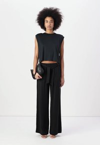 Black cropped sleeveless top paired with high-waisted black wide-leg trousers, featuring a tied waist. Model holds a small black handbag.