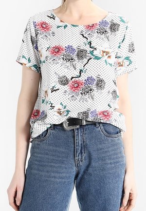 Kurzarm-T-Shirt mit weißem Hintergrund, schwarzen Punkten und bunten Blumenmustern. Kombiniert mit hochgeschnittenen blauen Jeans.