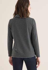 Vrouw met schouderlang bruin haar, gekleed in een zwart en grijs horizontaal gestreepte trui met lange mouwen en donkere broek, staande met haar rug naar voren.