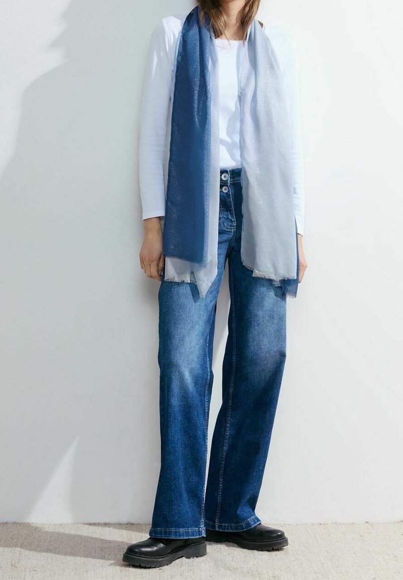 Personne portant une chemise blanche à manches longues, un jean bleu à jambes larges, des chaussures noires et une longue écharpe bleu bicolore devant un mur uni.