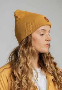 Żó�łta dzianinowa czapka z założonym mankietem i okrągłą skórzaną łatą. Modelka ma długie, falujące włosy, nosi lekką kurtkę i koszulkę.