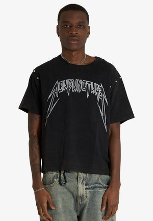 Giovane uomo che indossa una t-shirt nera oversize con la scritta "ACUPUNCTURE" in lettere bianche stilizzate e jeans chiari con portachiavi.