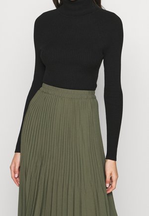 Kobieta ubrana w dopasowany czarny prążkowany golf i oliwkowo-zieloną plisowaną spódnicę, widoczna od szyi do talii.