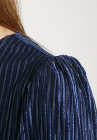 Marinblus med textur och vertikala veck samt puffärmar, med en metallisk lyster och mjukt tyg. Närbild av axelområdet.