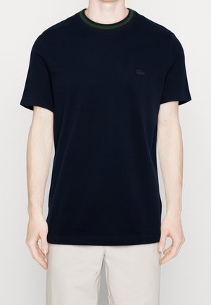 Homme portant un t-shirt uni bleu marine foncé à manches courtes avec un col rayé vert et rouge et un pantalon beige clair sur un fond uni.