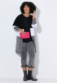 Eine Frau lächelt selbstbewusst in einem farblich blockierten Cardigan, schwarzem Oberteil, grauen fransigen Jeans, mit genagelten schwarzen Stiefeln und hält eine leuchtend pinke gesteppte Clutch.