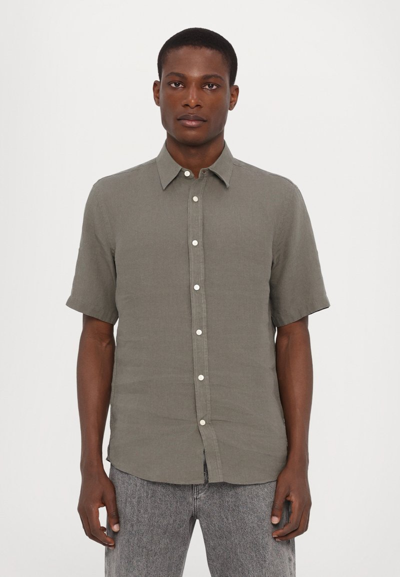 Korte mouwen shirt in olijfgroen, gemaakt van textuurstof, met een puntige kraag en een voorknoopsluiting. Draagt bij grijze jeans.